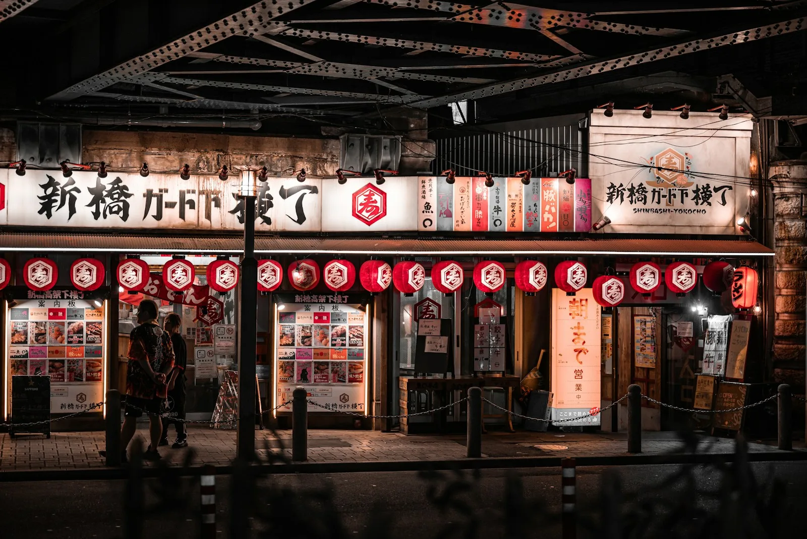 Façade de restaurants sous une voie ferrée la nuit, avec lanternes rouges, enseignes en kanji et vitrines de menus illustrés